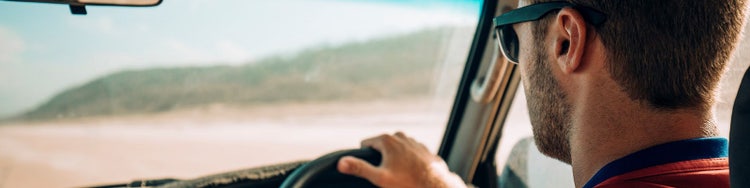 Man seated in the driver’s seat of a car, looking out through the open window towards a sandy landscape.