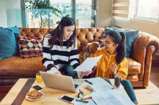 Two young women reviewing policy documents together in a cozy living room with a laptop and papers nearby